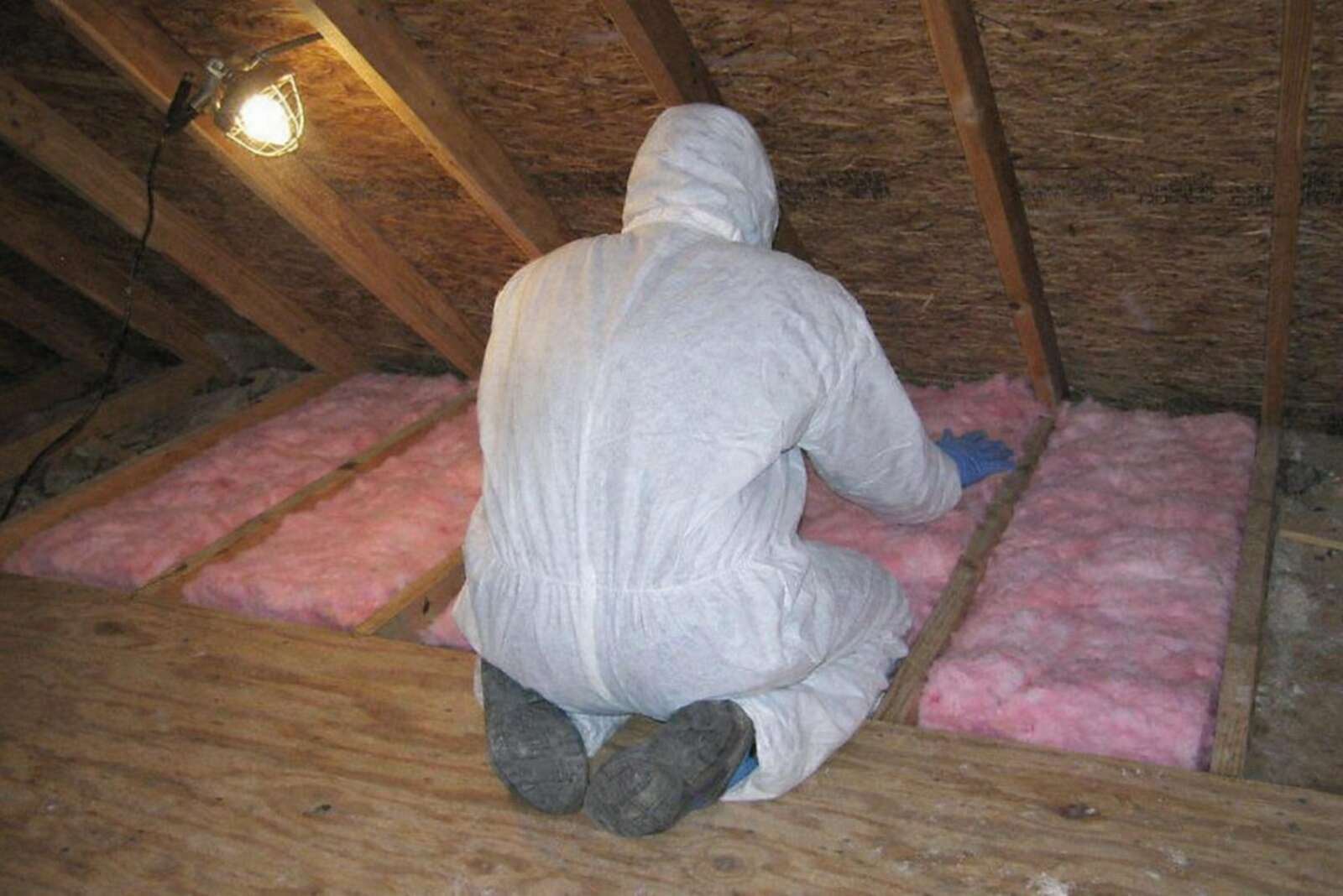 A gloved worker crouched beside pink fiberglass insulation, face out of frame