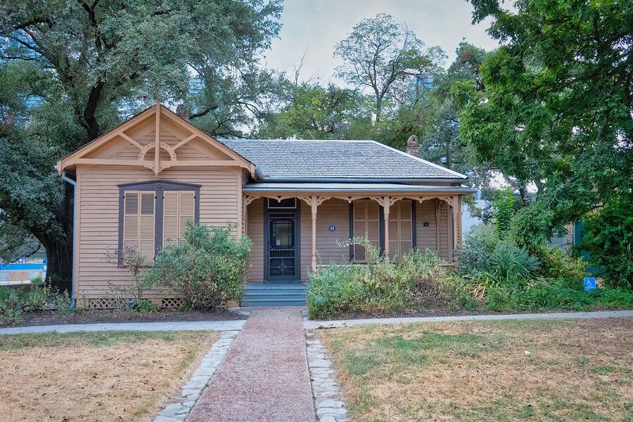 An older wood-frame cottage with a covered porch, set among trees in Texas